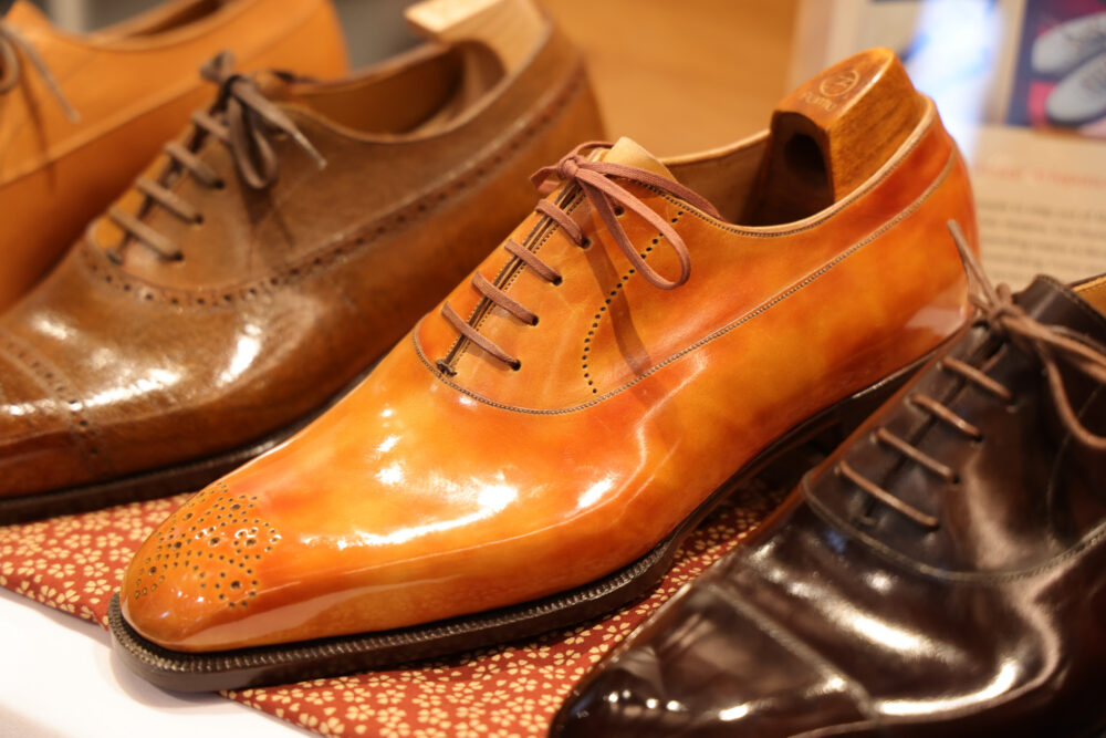 A polished orange leather dress shoe with brown laces, displayed alongside brown and black shoes.