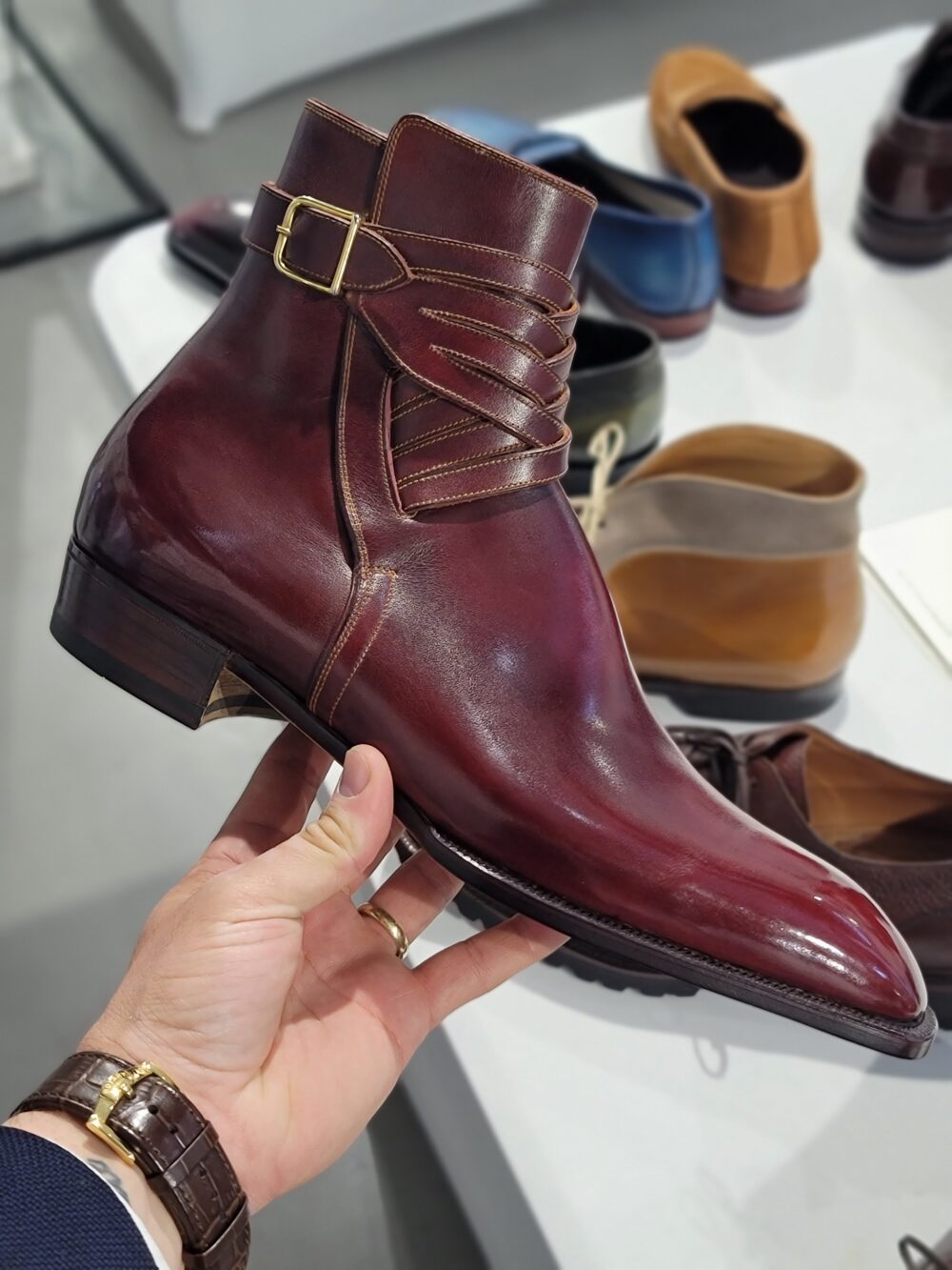 A person holds a polished burgundy leather ankle boot with a decorative strap and gold buckle.