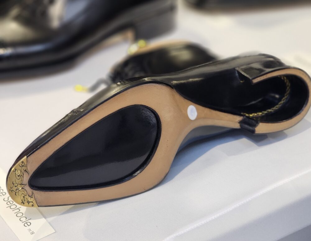 A black high-heeled shoe with a beige sole and decorative detailing on the heel.