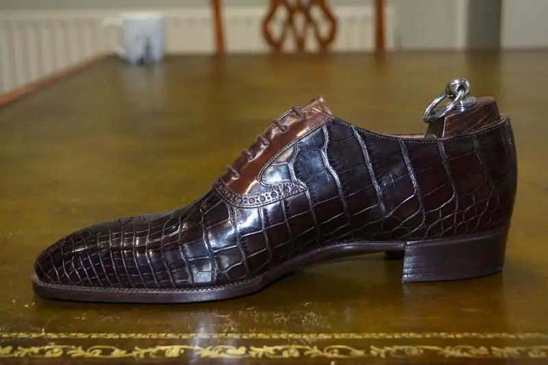 Luxurious men's dress shoe made from dark brown crocodile leather, featuring a classic lace-up design and elegant brogue detailing, displayed on a polished wooden table.