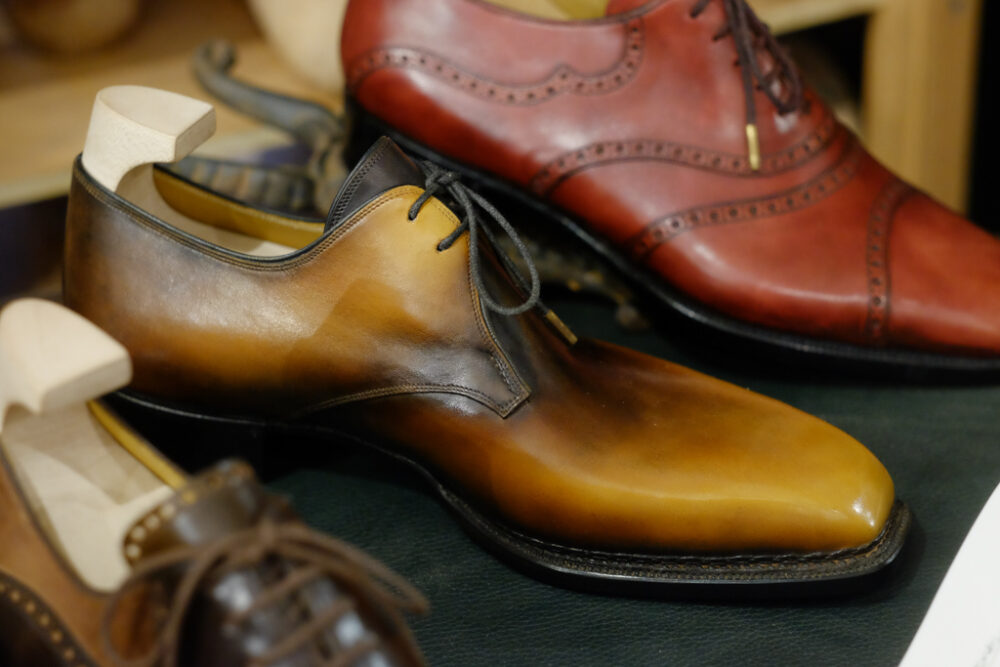 A polished brown leather shoe with a gradient finish, accompanied by a wooden shoe tree.