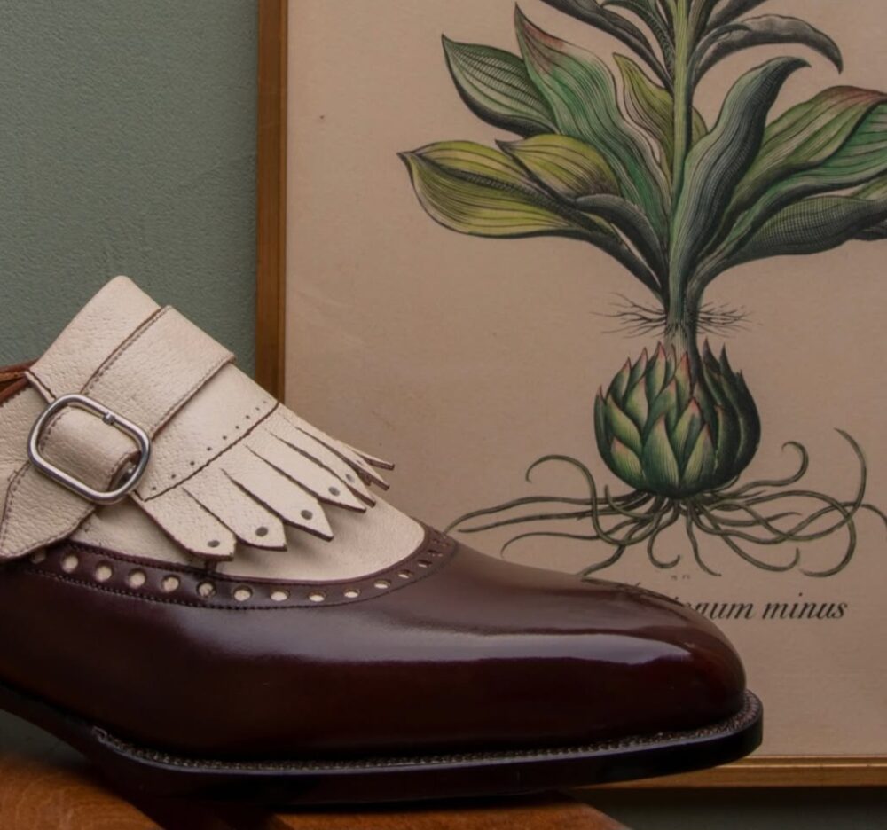 A brown leather shoe with a white fringed strap and a silver buckle.