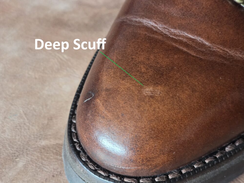 A brown leather shoe with a deep scuff marked by a green line on the toe.