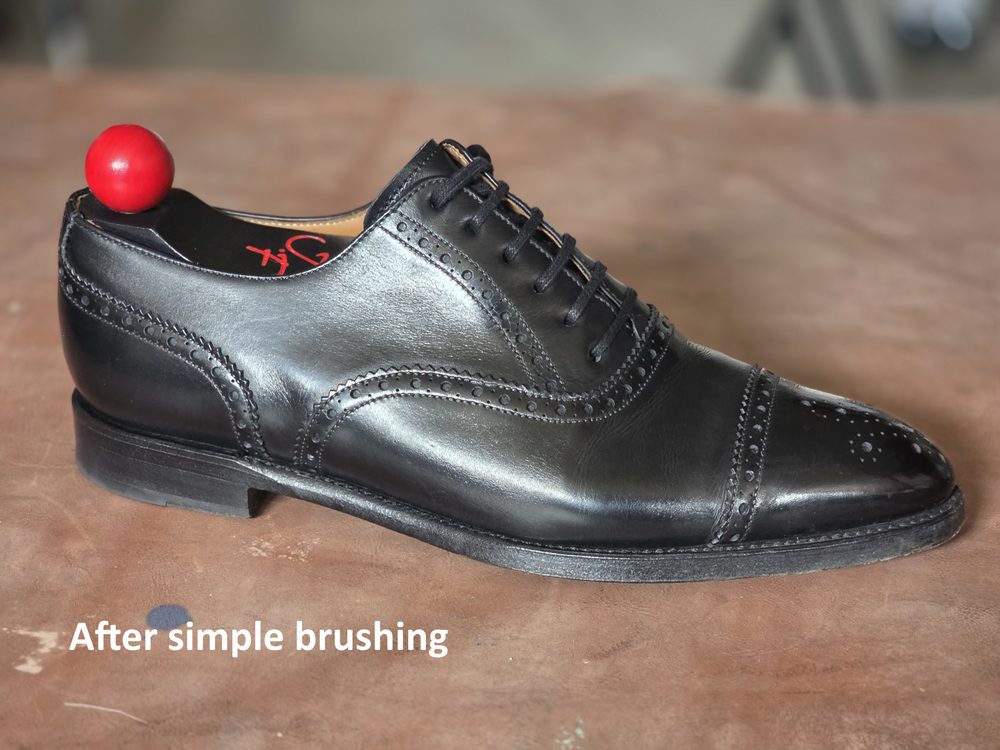 A polished black leather shoe with decorative perforations and a red shoehorn on the heel, labeled "After simple brushing."