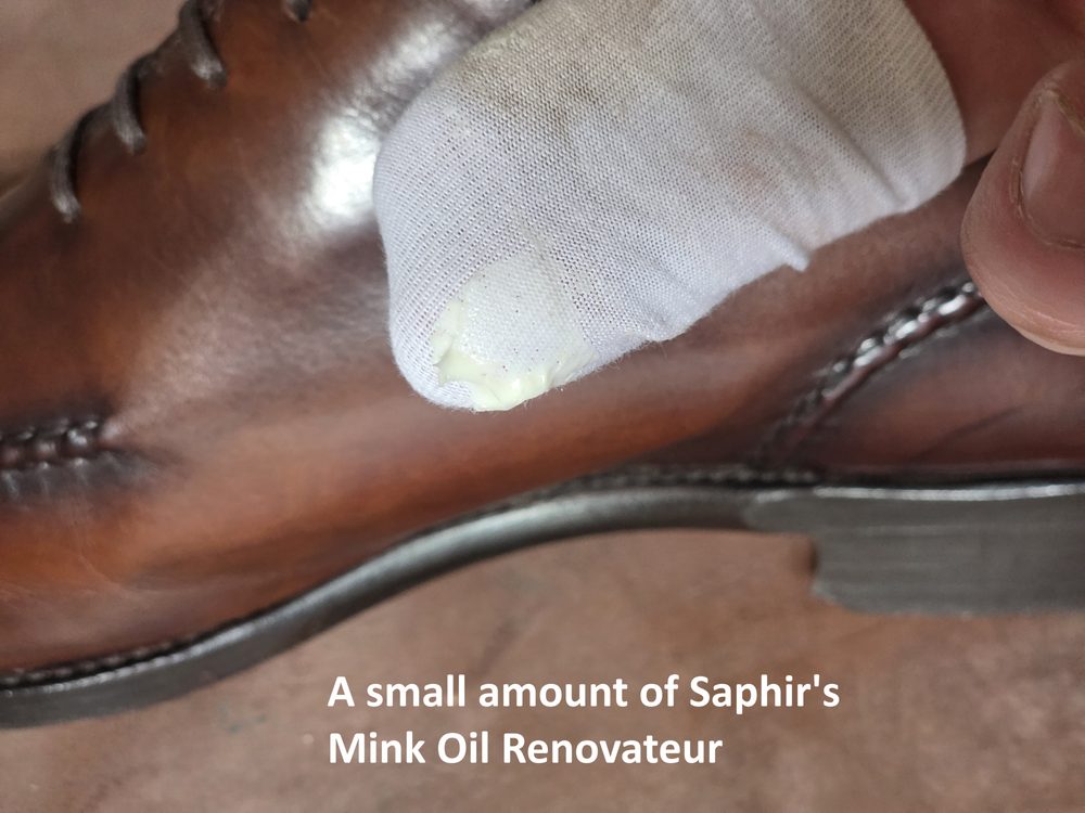 A close-up of a brown leather shoe with a finger applying Saphir's Mink Oil Renovateur.