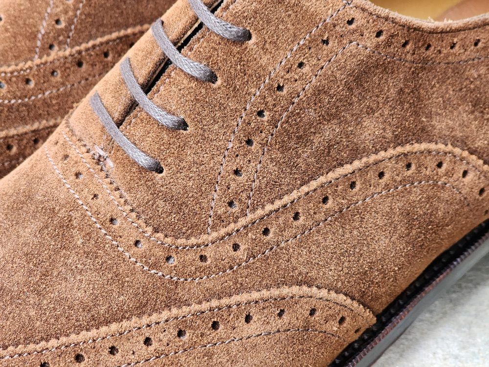Close-up of a brown suede brogue shoe featuring decorative perforations and gray laces.