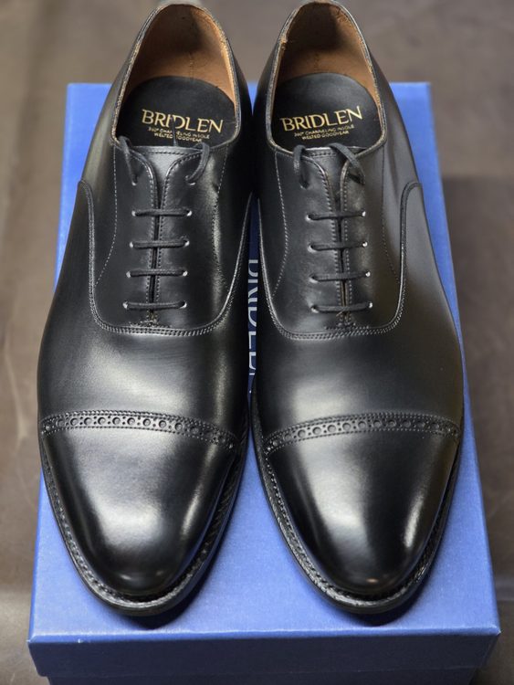 A pair of black leather Oxford shoes with decorative perforations, displayed on a blue box.