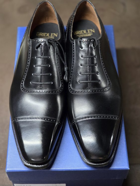 A pair of black leather dress shoes with a cap toe design, displayed on a blue box.