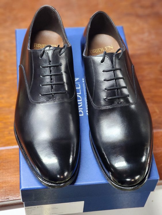A pair of black leather dress shoes with a sleek design and lace-up closure, placed on a blue box.