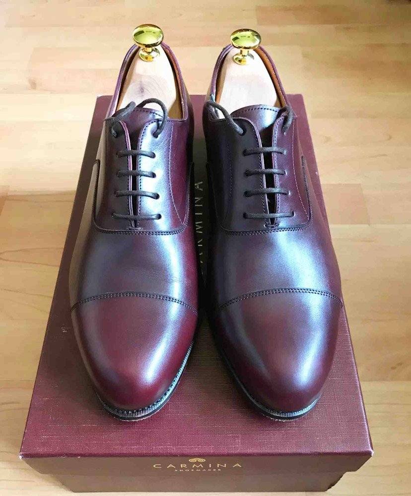 A pair of polished burgundy leather dress shoes with black laces, resting on a maroon shoe box.