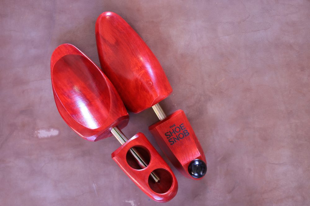 A pair of red wooden shoe trees with a metallic rod, featuring "THE SHOE SNOB" branding on one.
