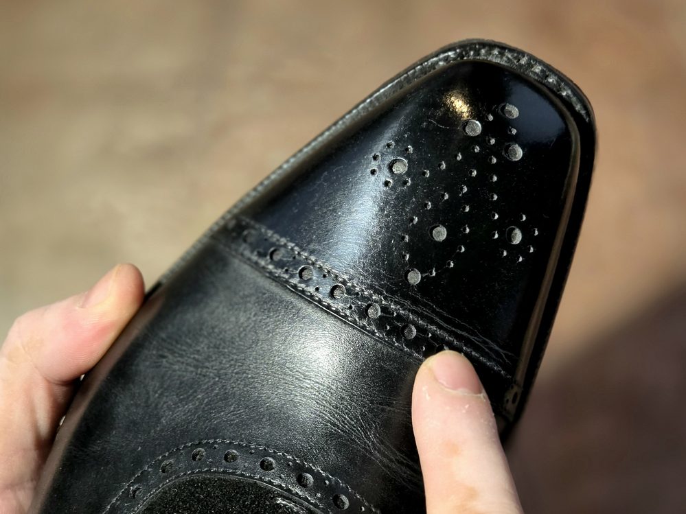 A close-up of a black leather shoe being held, showing a polished toe with visible smudges, illustrating a common shoe shine don't.