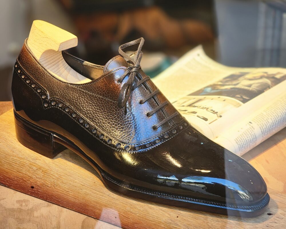 A polished black leather shoe with a textured brown panel and wooden shoe tree, illustrating Shoe Industry Lies.