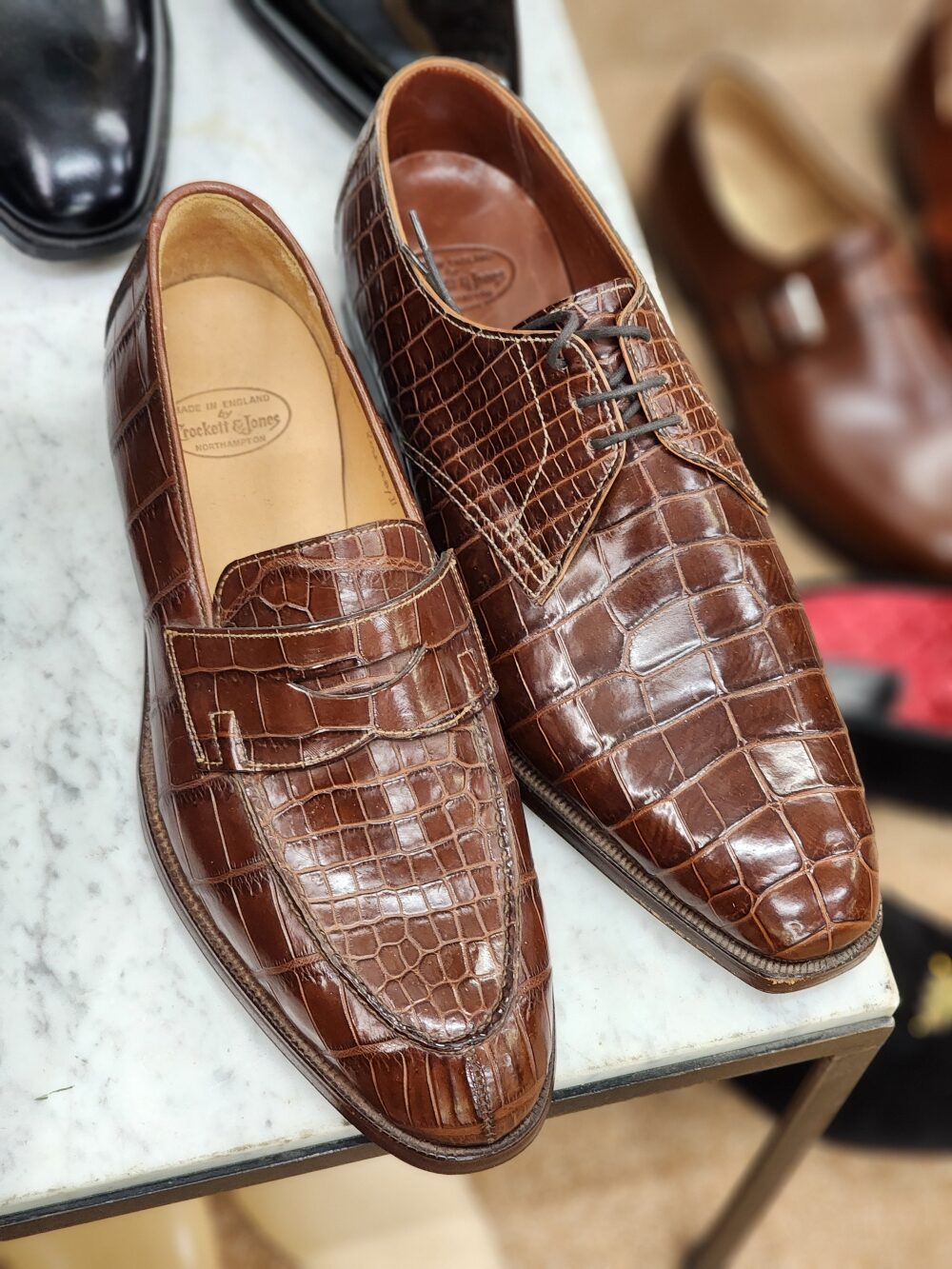 A pair of brown crocodile leather shoes, one a loafer and the other a lace-up style, displayed on a marble surface.