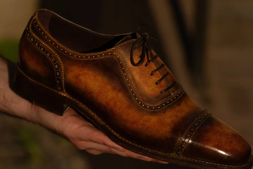 A hand holds a polished brown leather dress shoe with decorative stitching and laces.