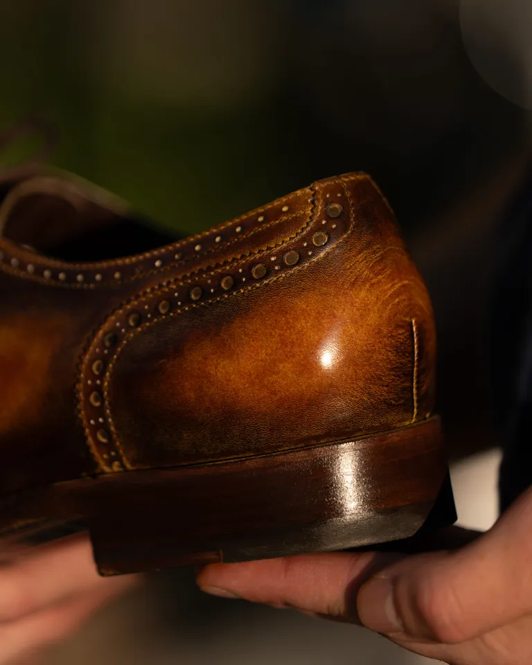 A close-up of a brown leather dress shoe being held, featuring decorative stitching along the edges.
