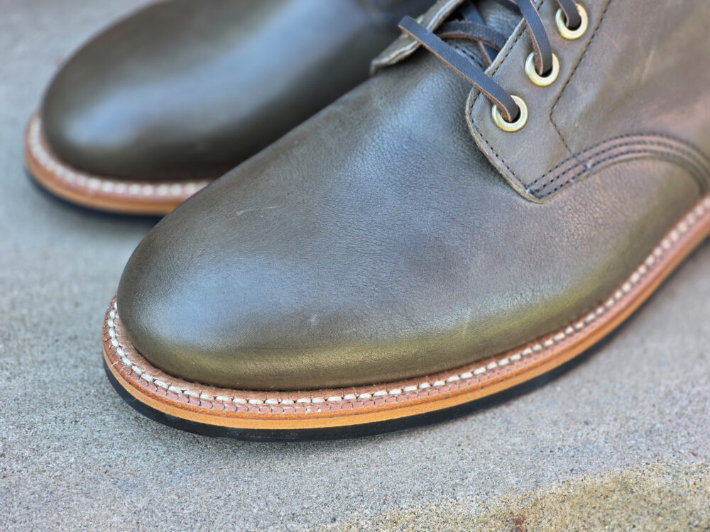 A close-up of two dark green leather shoes with brown soles and beige stitching.