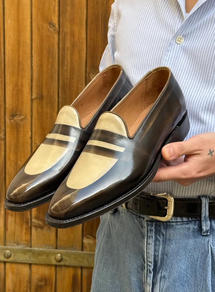 A pair of glossy black and beige leather loafers with striped detailing, held by a person.