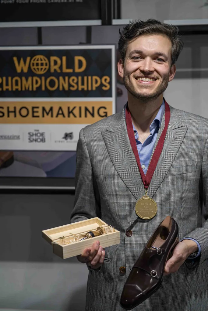 A smiling man in a gray suit holds a brown shoe and a wooden box containing shoemaking tools, wearing a gold medal.