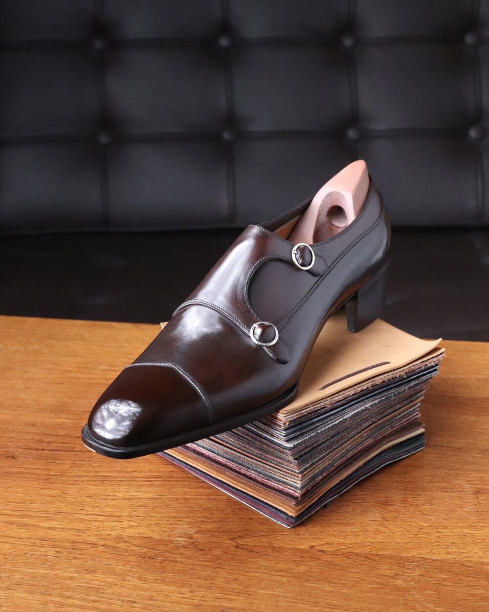 A polished brown leather shoe with a low heel and decorative buckles rests on a stack of colorful leather swatches.