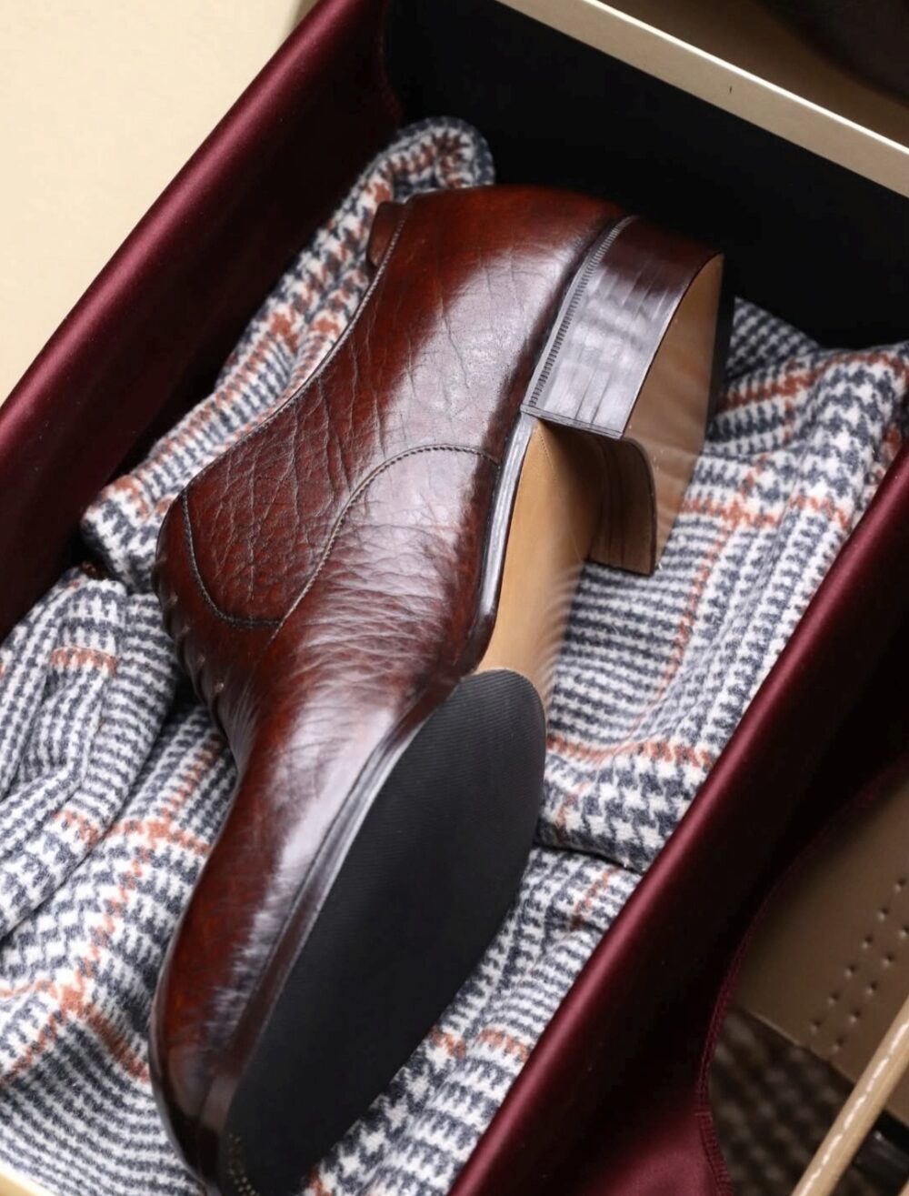 A polished brown leather shoe rests on a patterned cloth inside a box.