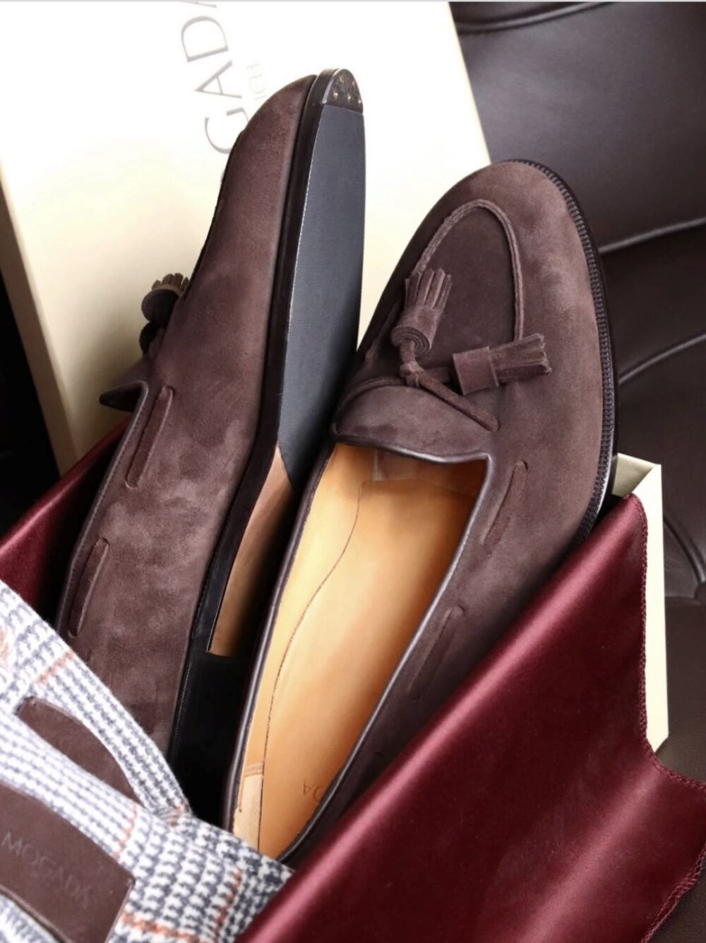 A pair of dark brown suede loafers with tassels, partially inside a bag.