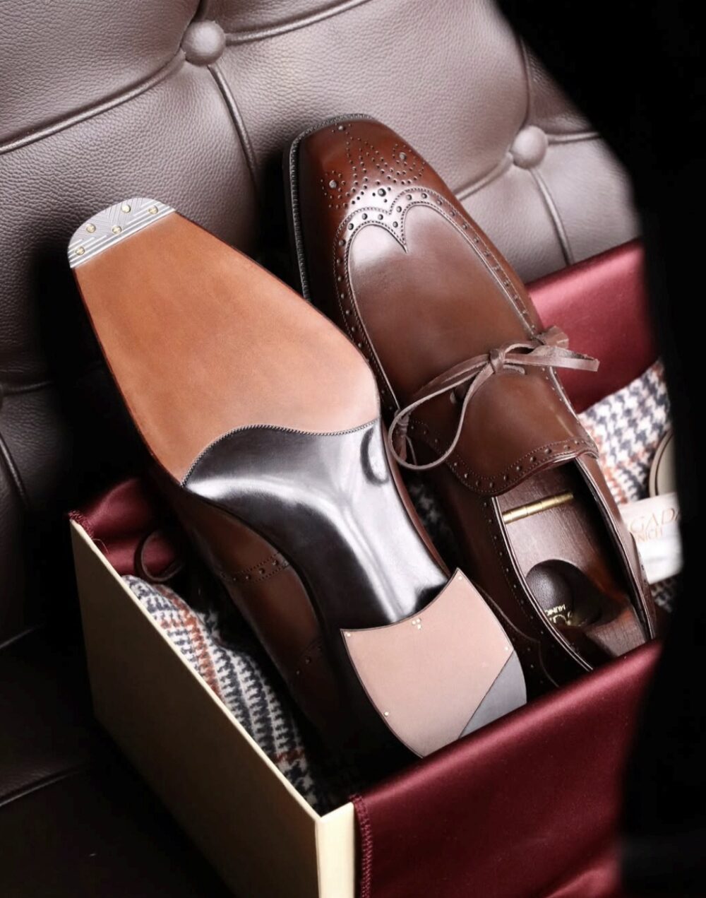 A pair of brown leather dress shoes with decorative perforations, one showing its smooth sole and the other with a leather cover, placed in a gift box.
