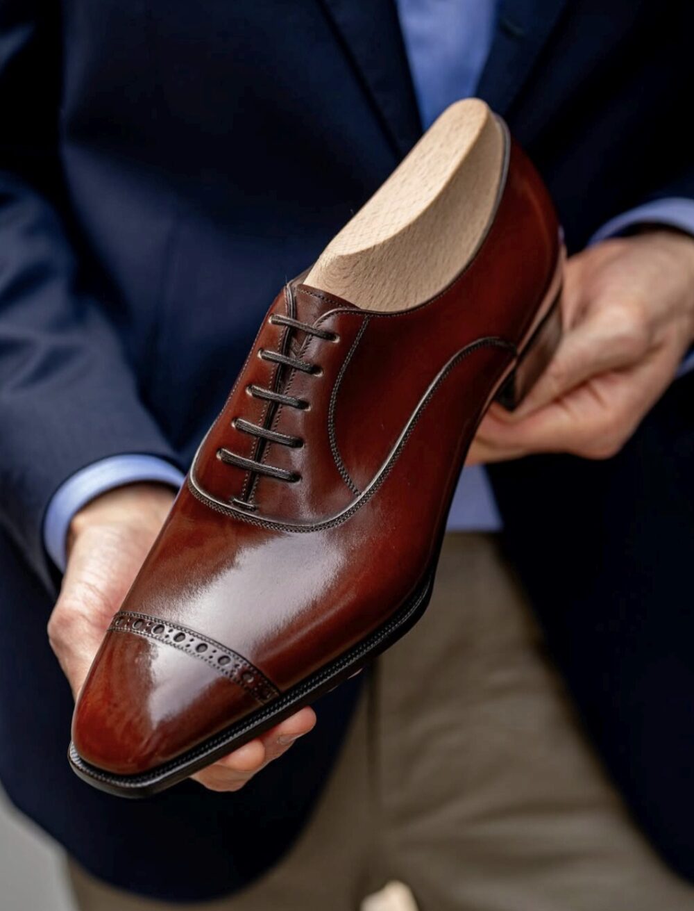 A polished brown leather dress shoe with decorative stitching and laces, held by a person wearing a dark suit.