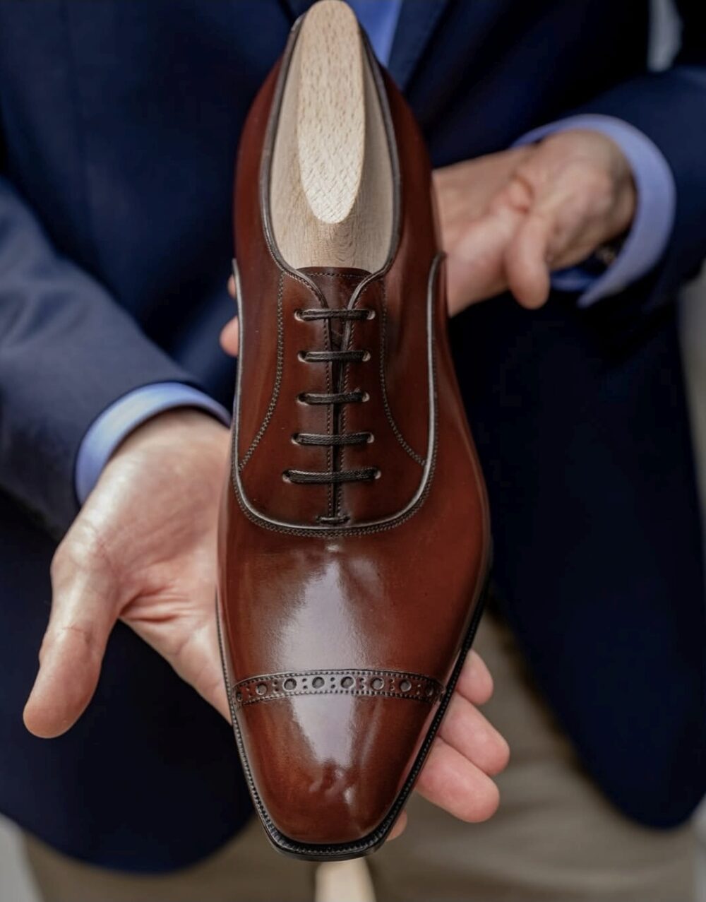 A person holds a polished brown leather dress shoe with a lace-up design and decorative perforations.