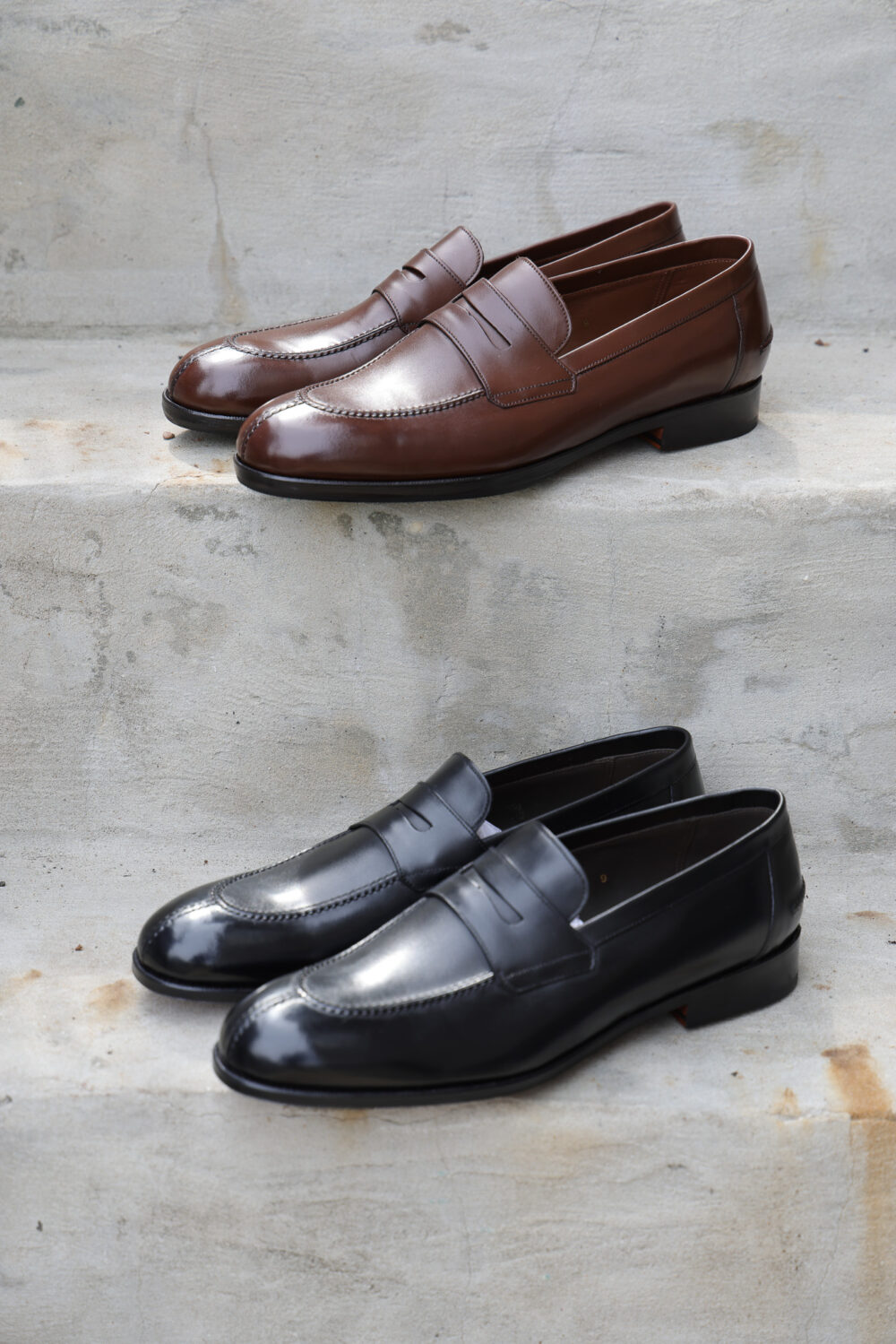 A pair of polished brown loafers sits above a pair of polished black loafers on concrete steps.