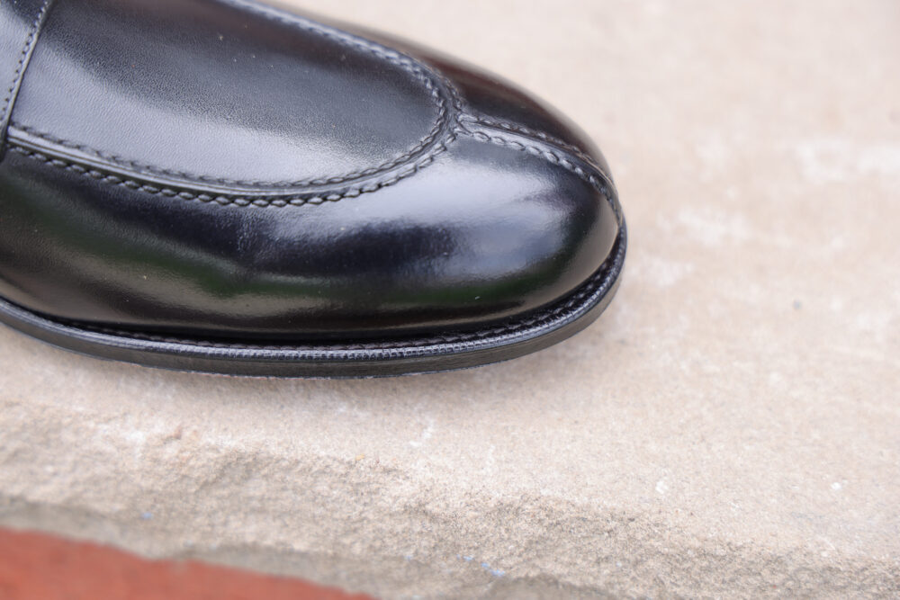 A close-up of a polished black leather shoe with a rounded toe and decorative stitching.