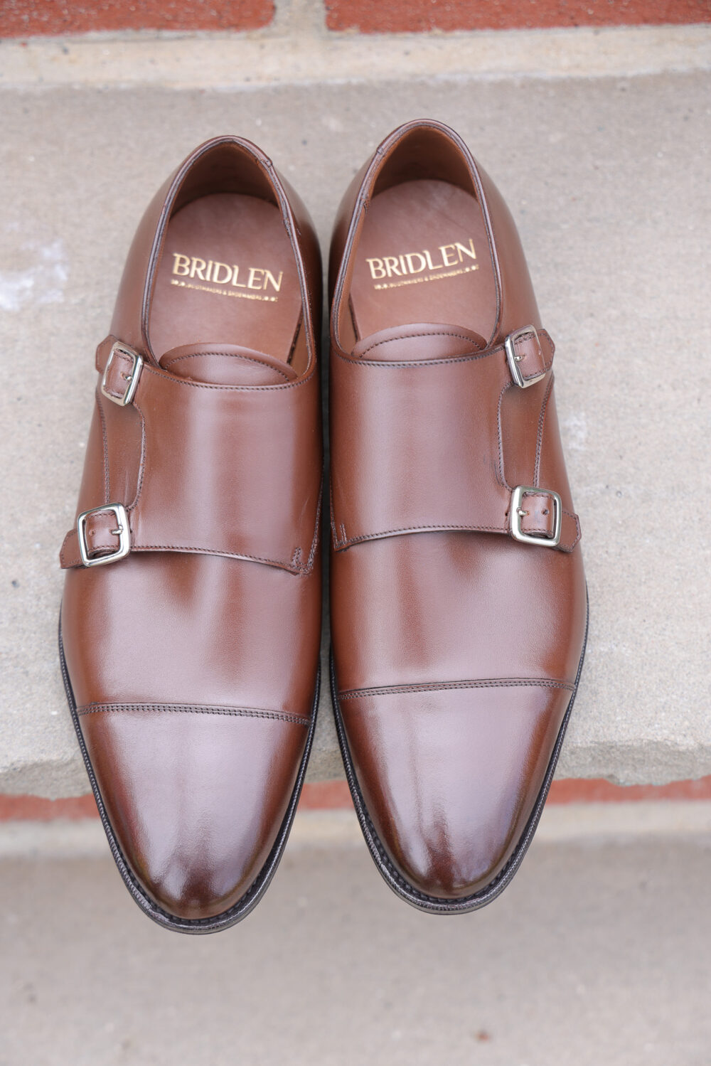 A pair of polished brown leather double monk strap shoes with silver buckles.