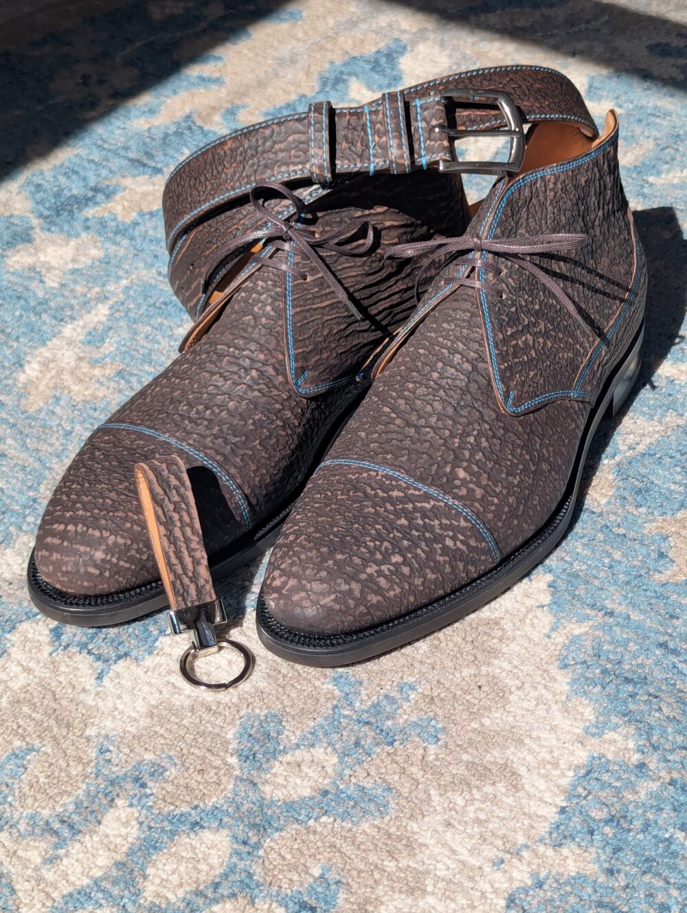 A pair of dark brown textured shoes with blue stitching and a matching belt.