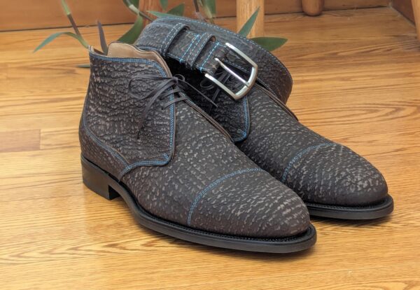 A pair of dark brown textured leather boots with blue stitching and a buckle strap.