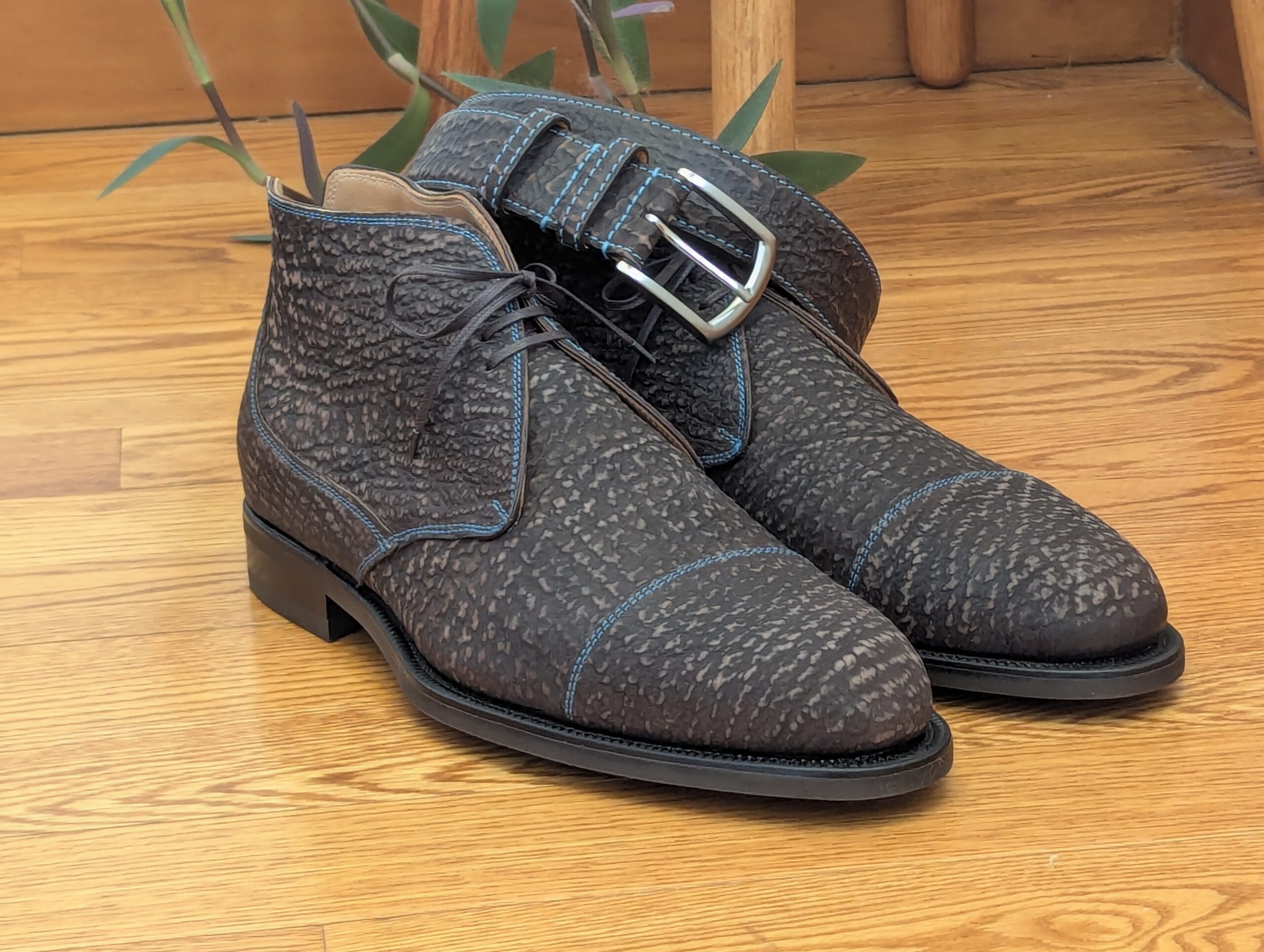 A pair of dark brown textured leather boots with blue stitching and a buckle strap.