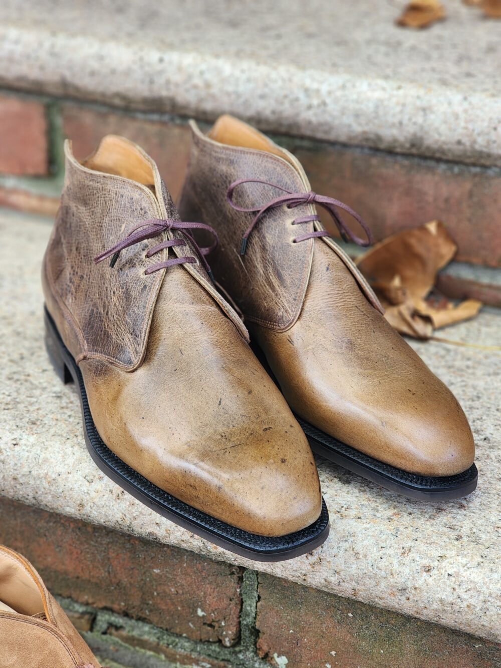 A pair of brown leather dress boots with purple laces, displayed on a stone surface.