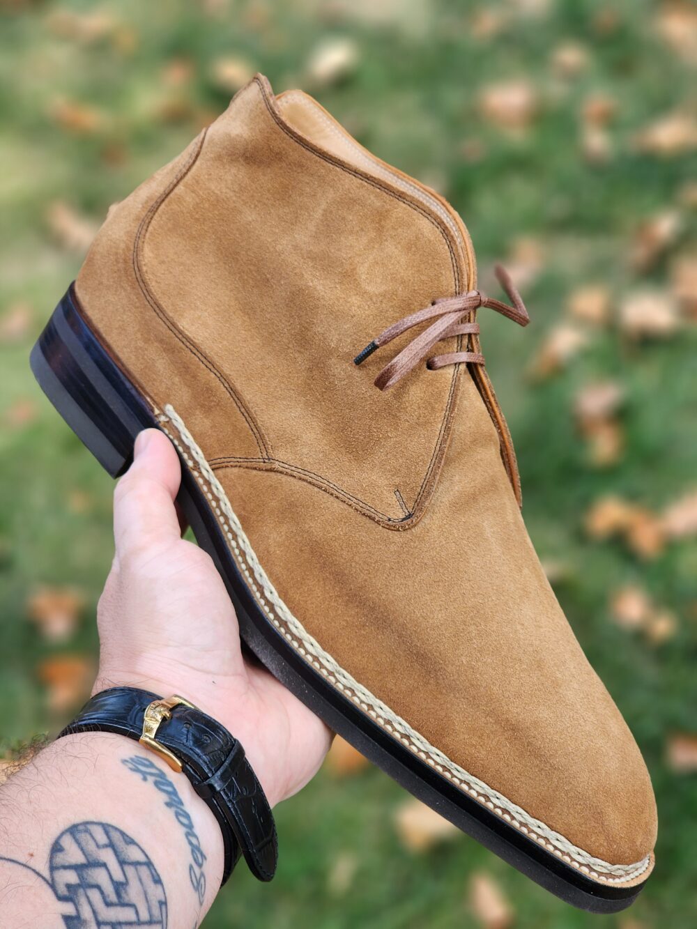 A hand holds a tan suede boot with brown laces and a braided sole.