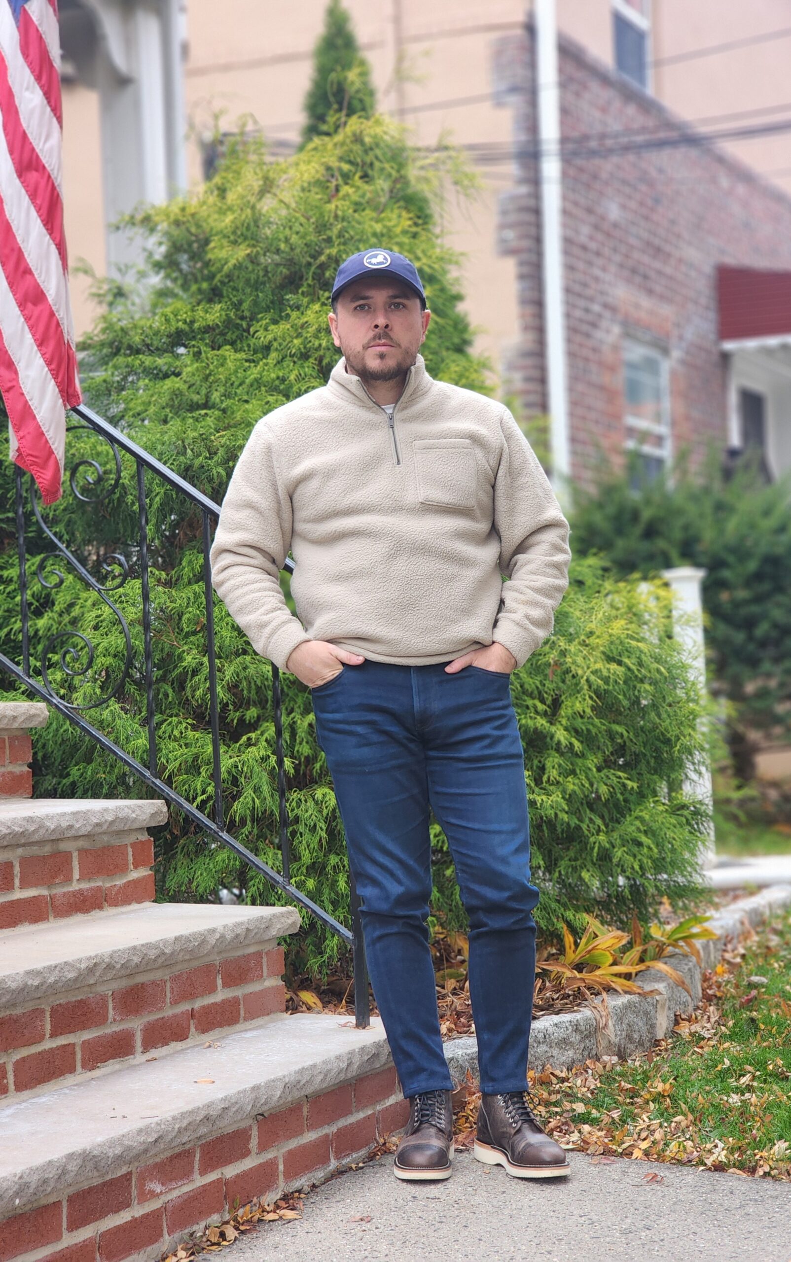A man stands on steps wearing a beige fleece pullover, blue jeans, and brown boots, with a navy cap and an American flag nearby.