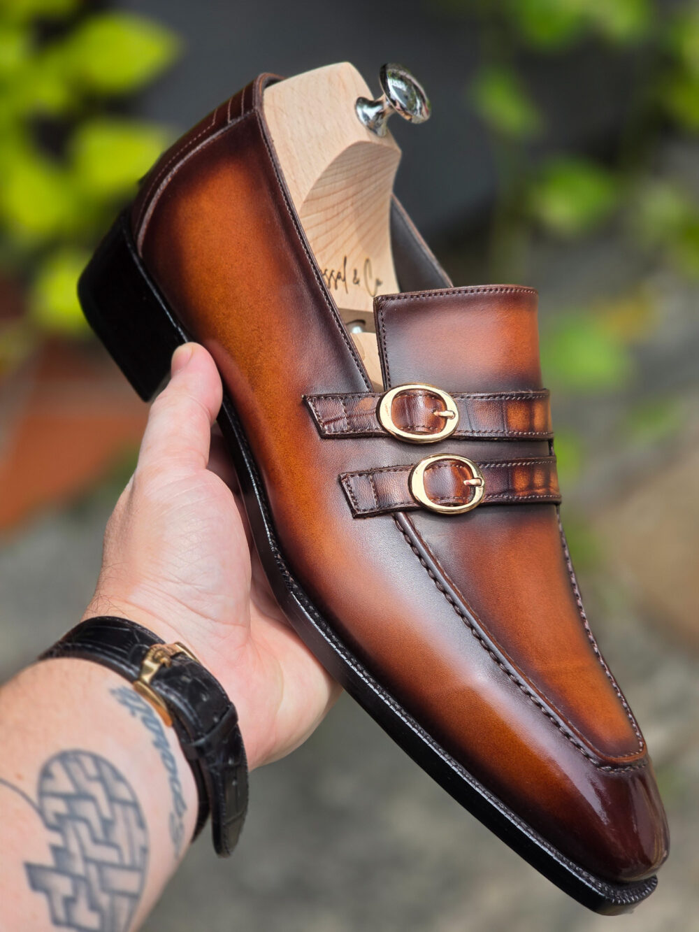 A hand holds a polished brown leather loafer with gold buckles 