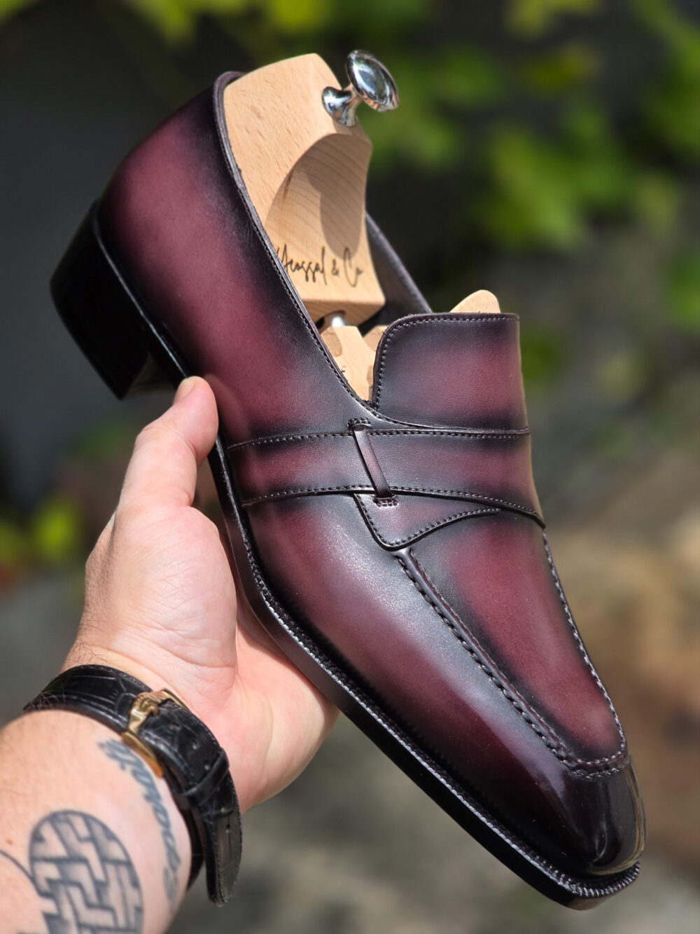 A hand holds a burgundy leather shoe with a butterfly strap design