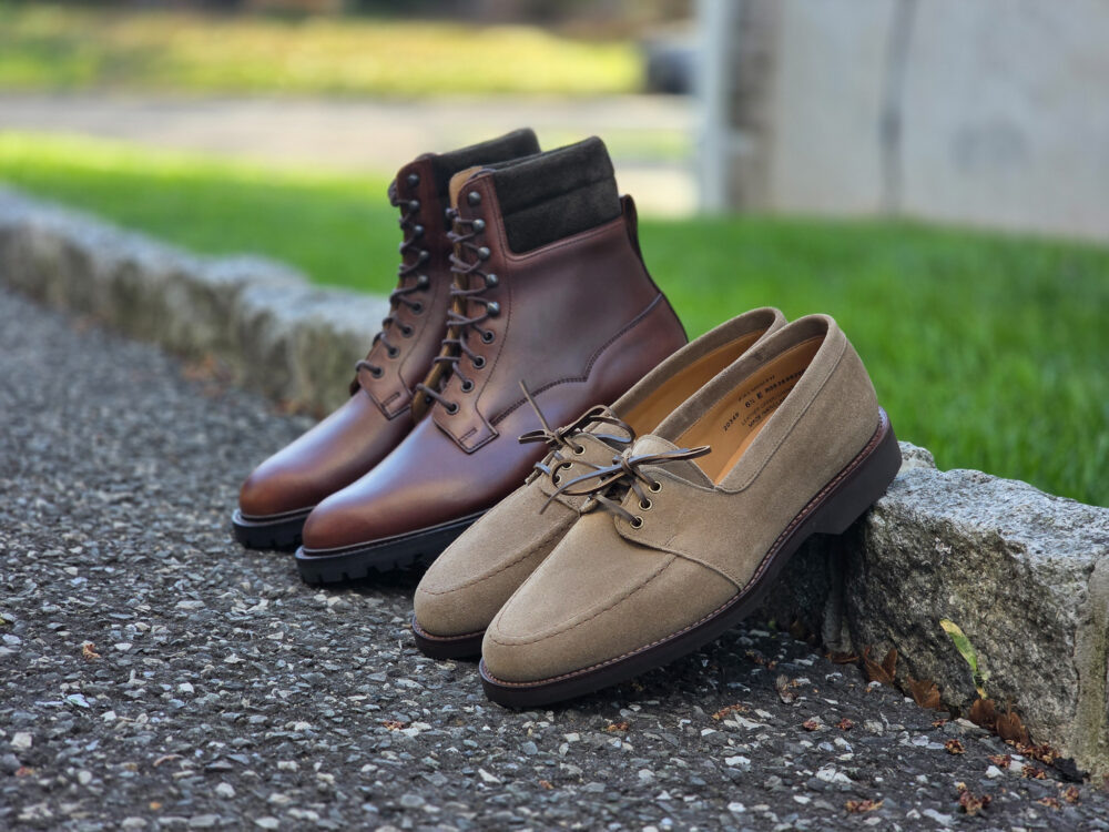 A pair of brown leather boots and a pair of beige suede shoes are placed side by side on a gravel surface.