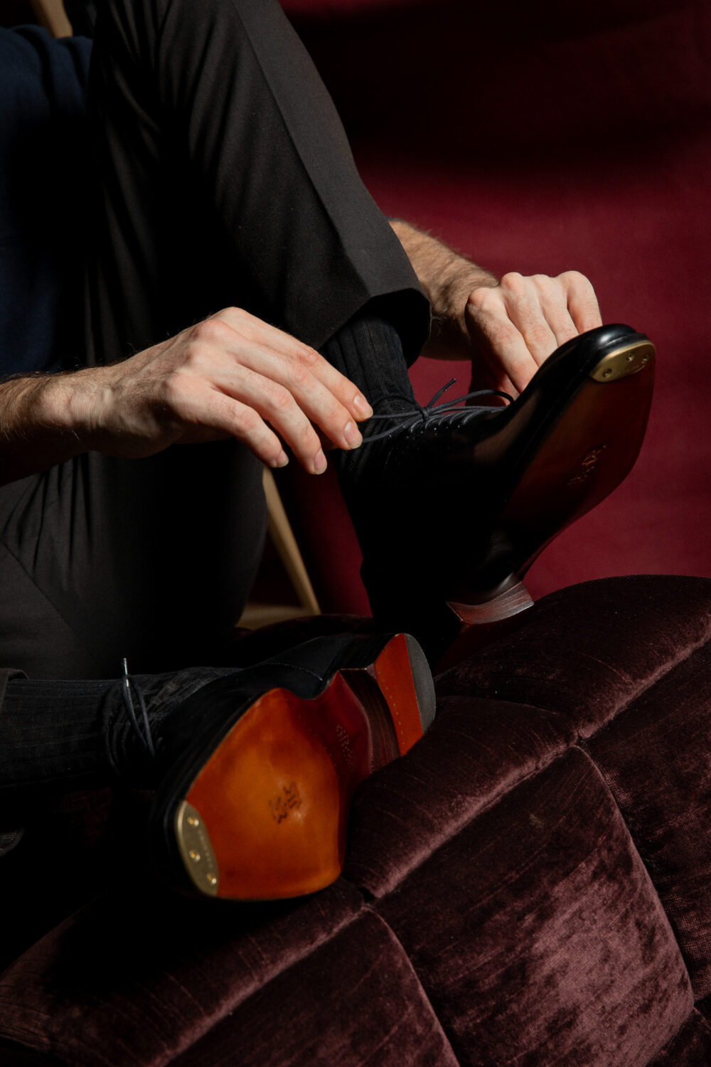 A person with light skin is tying a black leather dress shoe with a brown sole while sitting down.