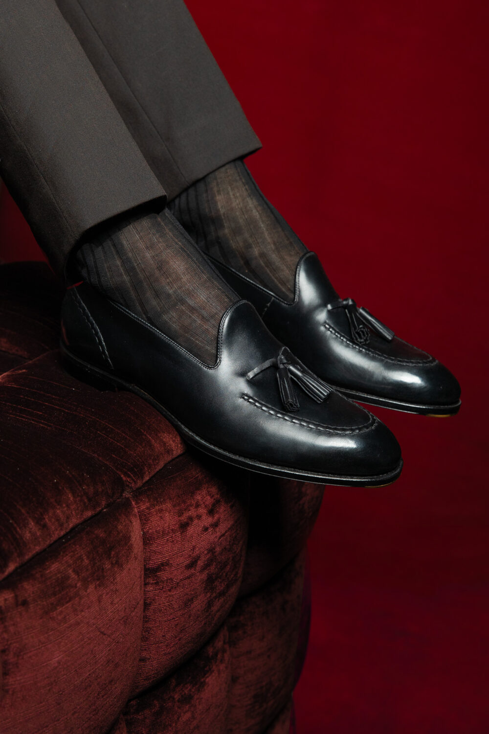 Black leather loafers with tassels, worn with sheer striped socks and brown trousers.