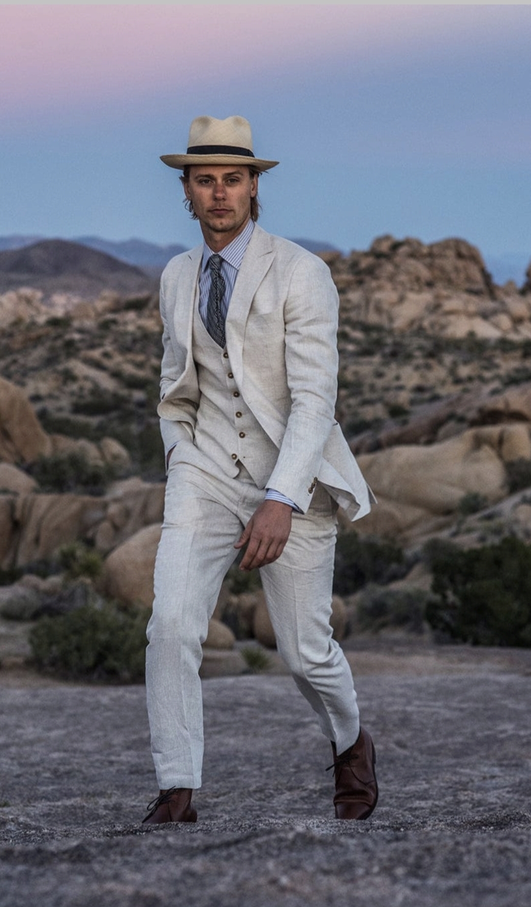 A man in a light-colored linen suit and straw hat walks confidently across rocky terrain.