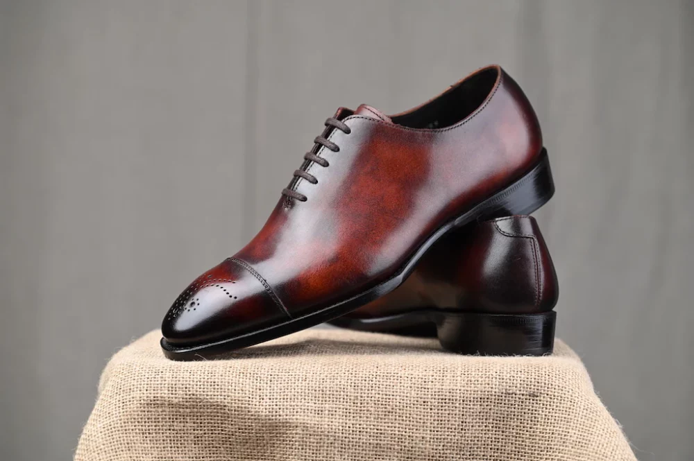 A pair of polished brown leather dress shoes with a cap toe design, displayed on a textured surface.