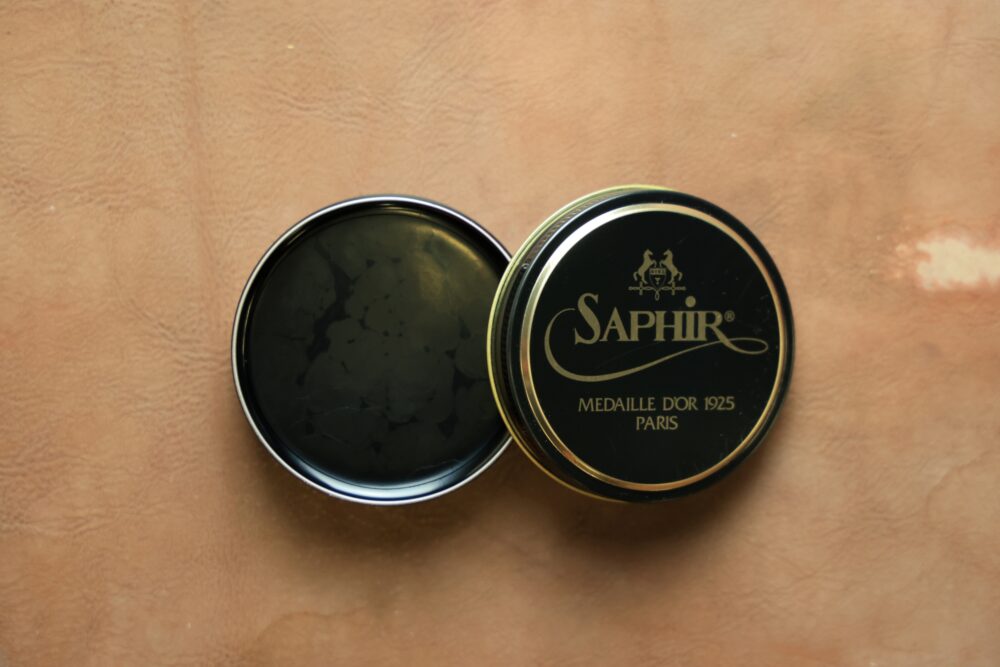 A black tin of Saphir shoe polish with a gold logo and text, opened to reveal the glossy polish inside.