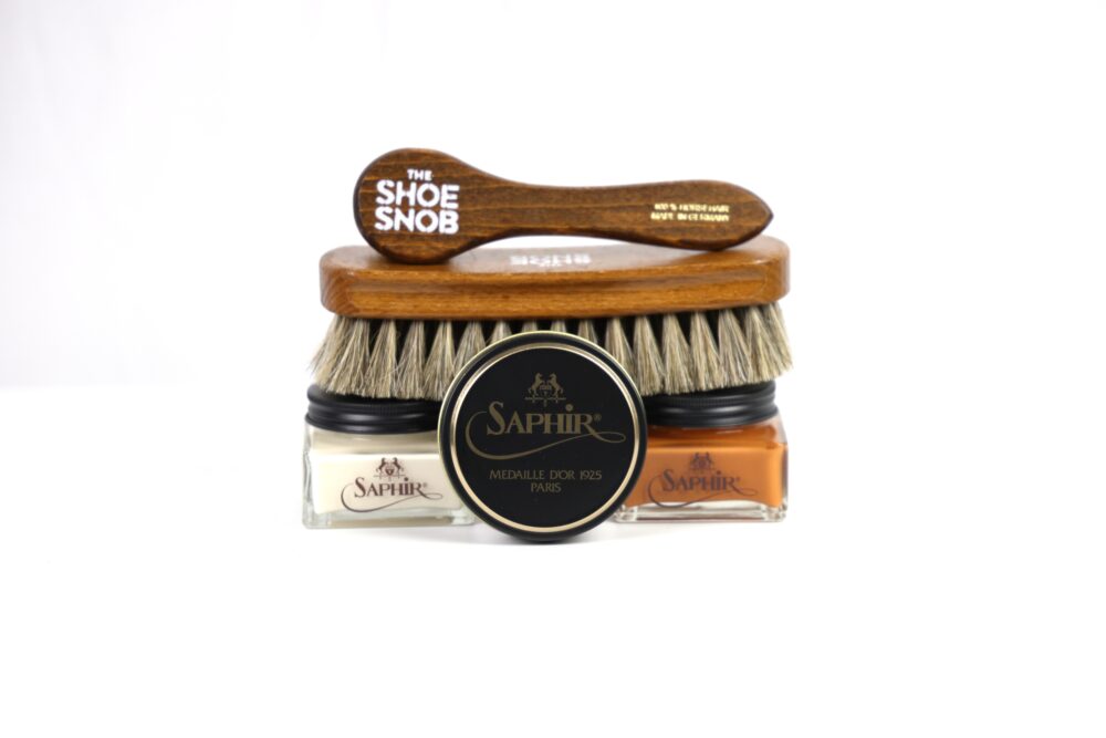 A wooden shoe brush labeled "The Shoe Snob" rests atop two jars of Saphir cream polish, one white and one brown, with a black Saphir polish tin in the center.