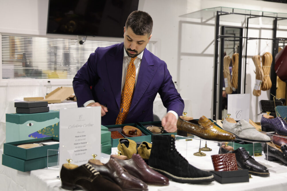 A man in a purple suit is arranging various styles of leather shoes and accessories on a table.