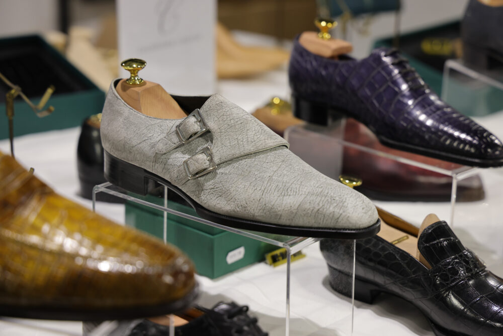 A pair of light gray leather shoes with double buckles displayed on a clear stand.