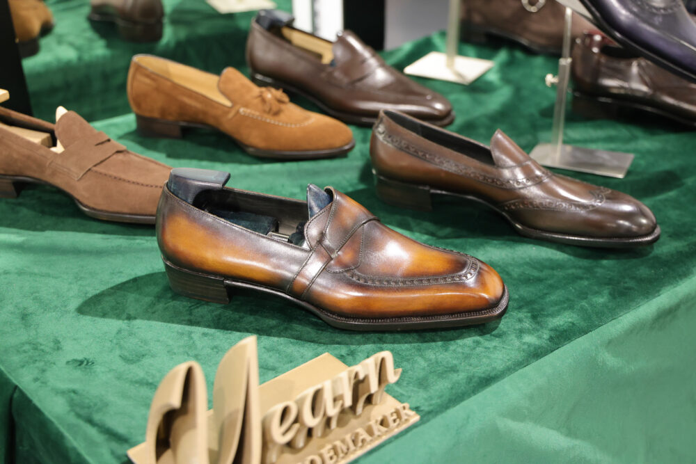 A polished brown leather shoe with a decorative seam is prominently displayed among other styles on a green surface.
